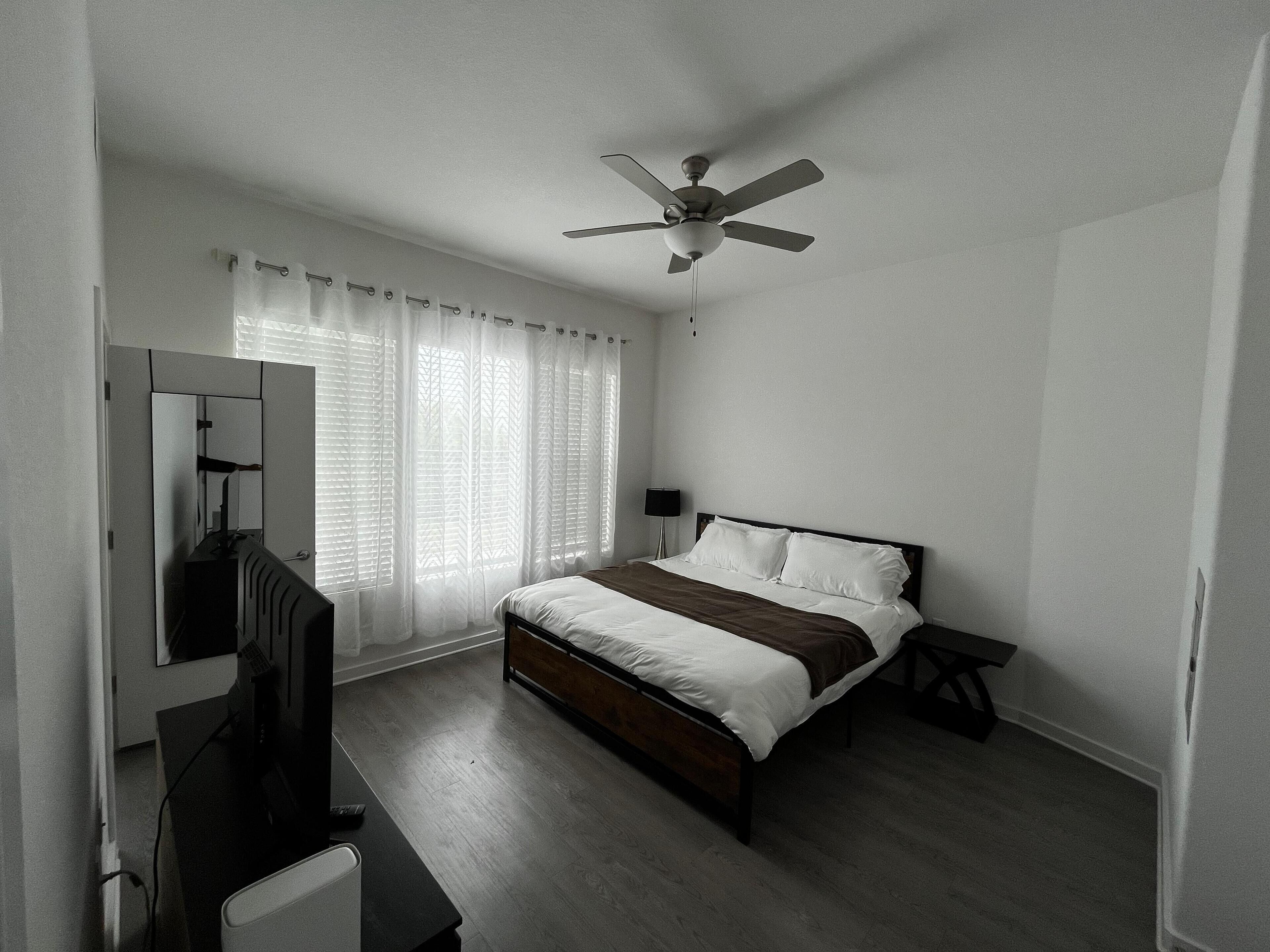 Ein Schlafzimmer mit einem Bett, Deckenventilator, Nachttisch, Lampe, Kommode und Spiegel.