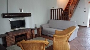 Een woonkamer met een schouw van baksteen, een grijze bank, rieten stoelen en een houten trap.