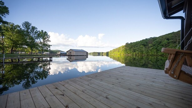A wooden dock extending over calm water, with a building in the distance and a forested hill.
