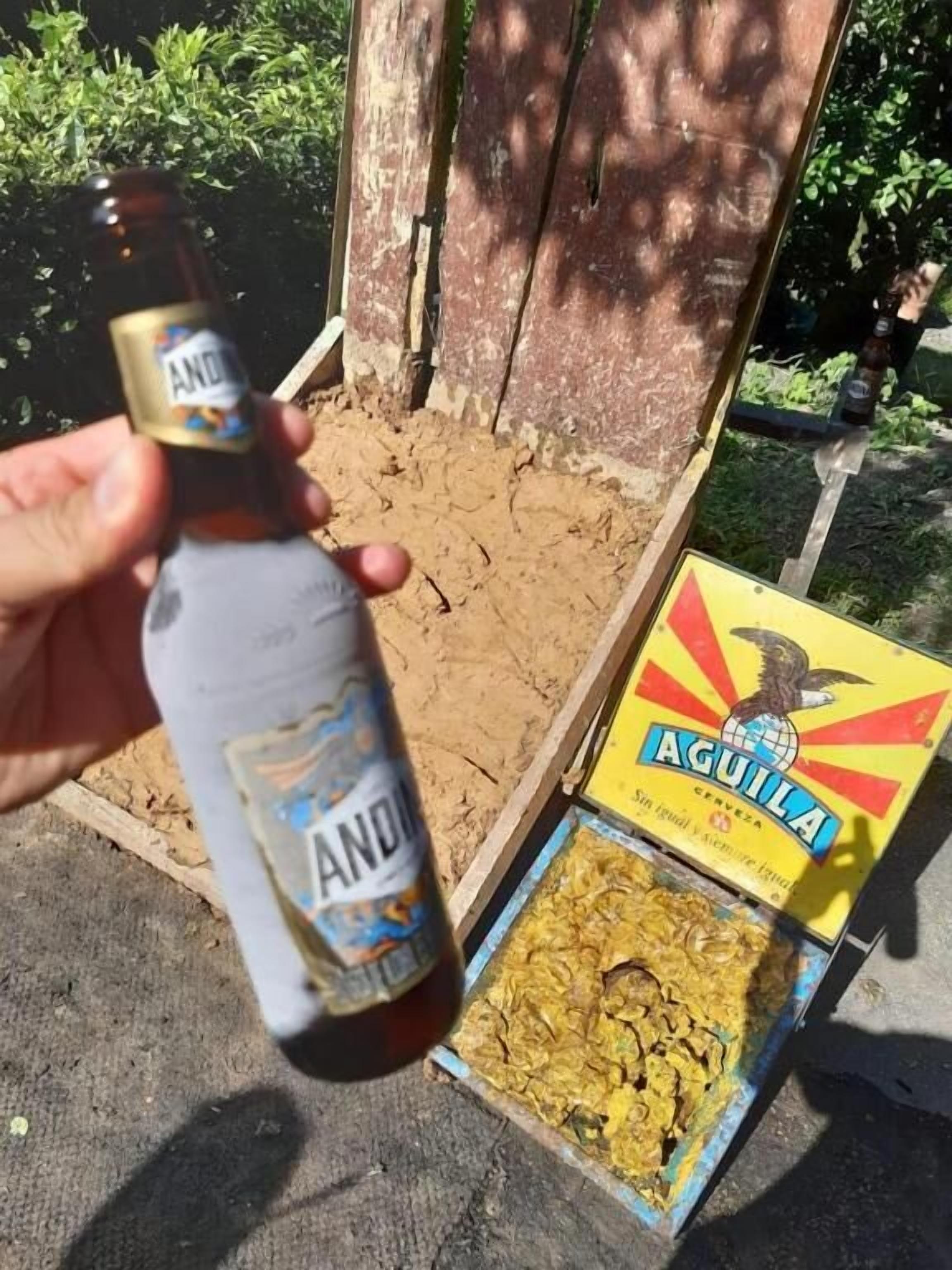 A hand holding a bottle of Andina beer, with a box of Aguila beer and a wooden crate in the background.