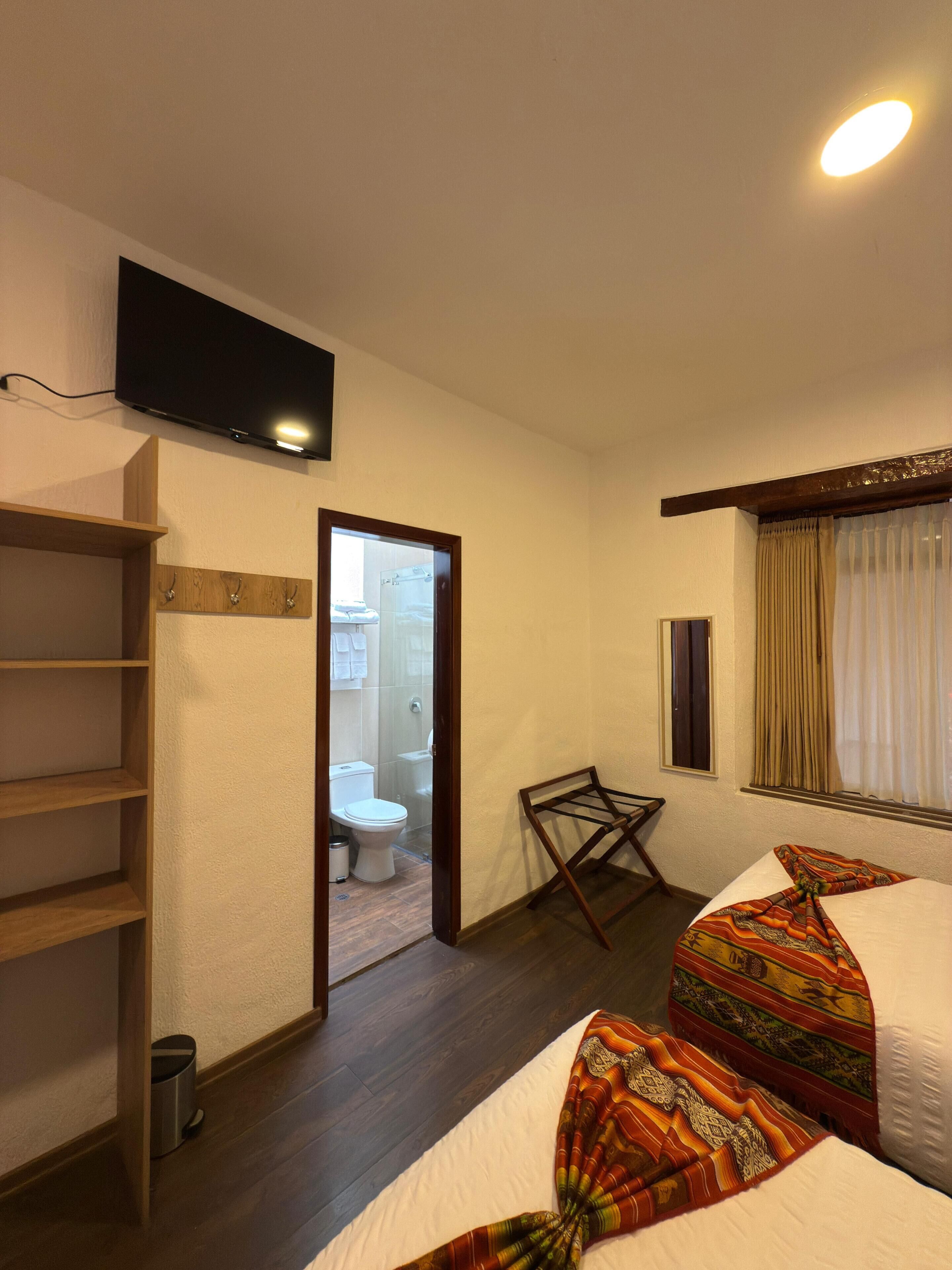 A hotel room with a bed, a TV mounted on the wall, a wooden shelf, and a window with curtains.