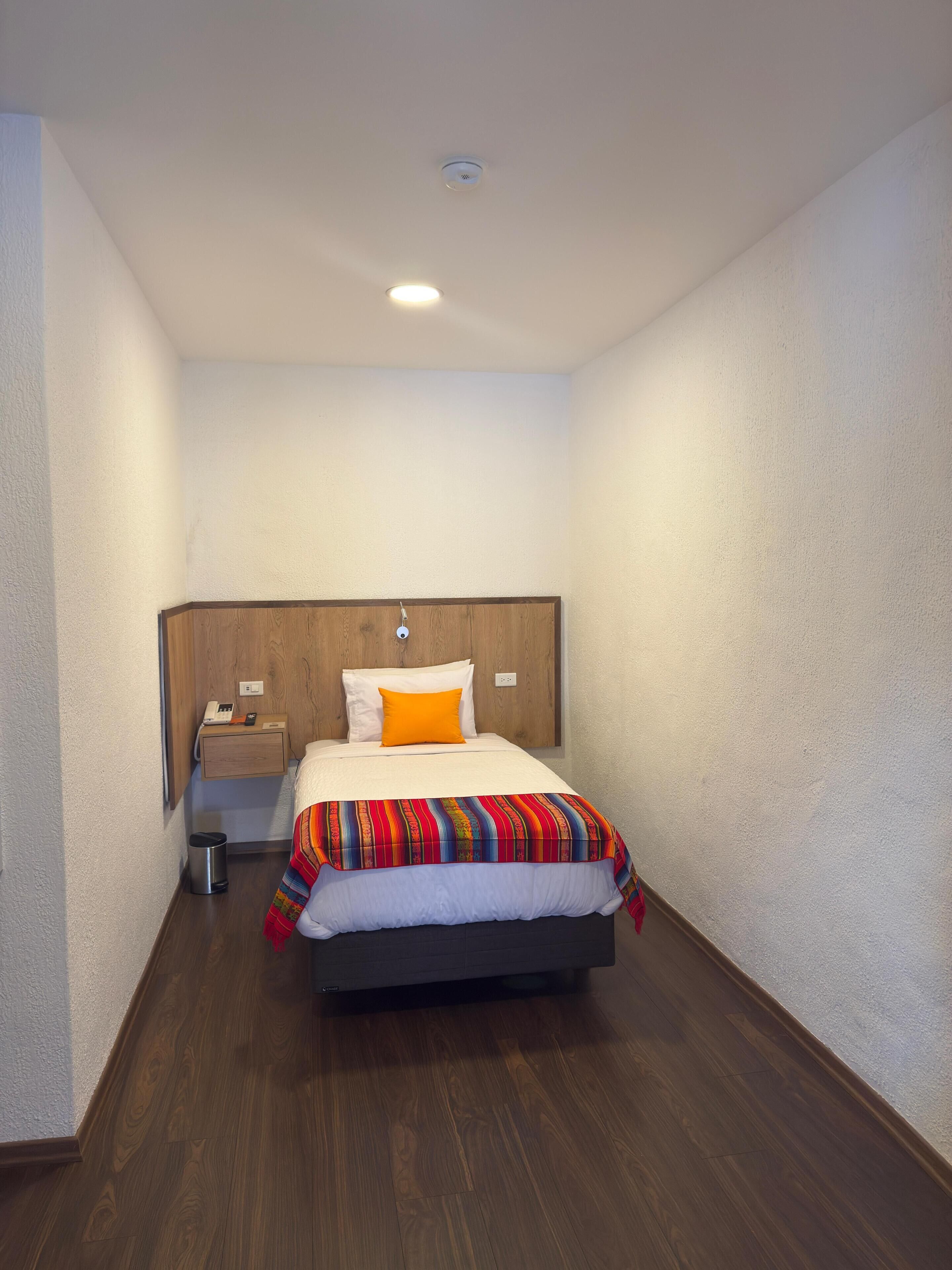 A hotel room with a wooden headboard, a single bed with an orange pillow, a bedside table with a phone and a lamp, and a small trash bin.
