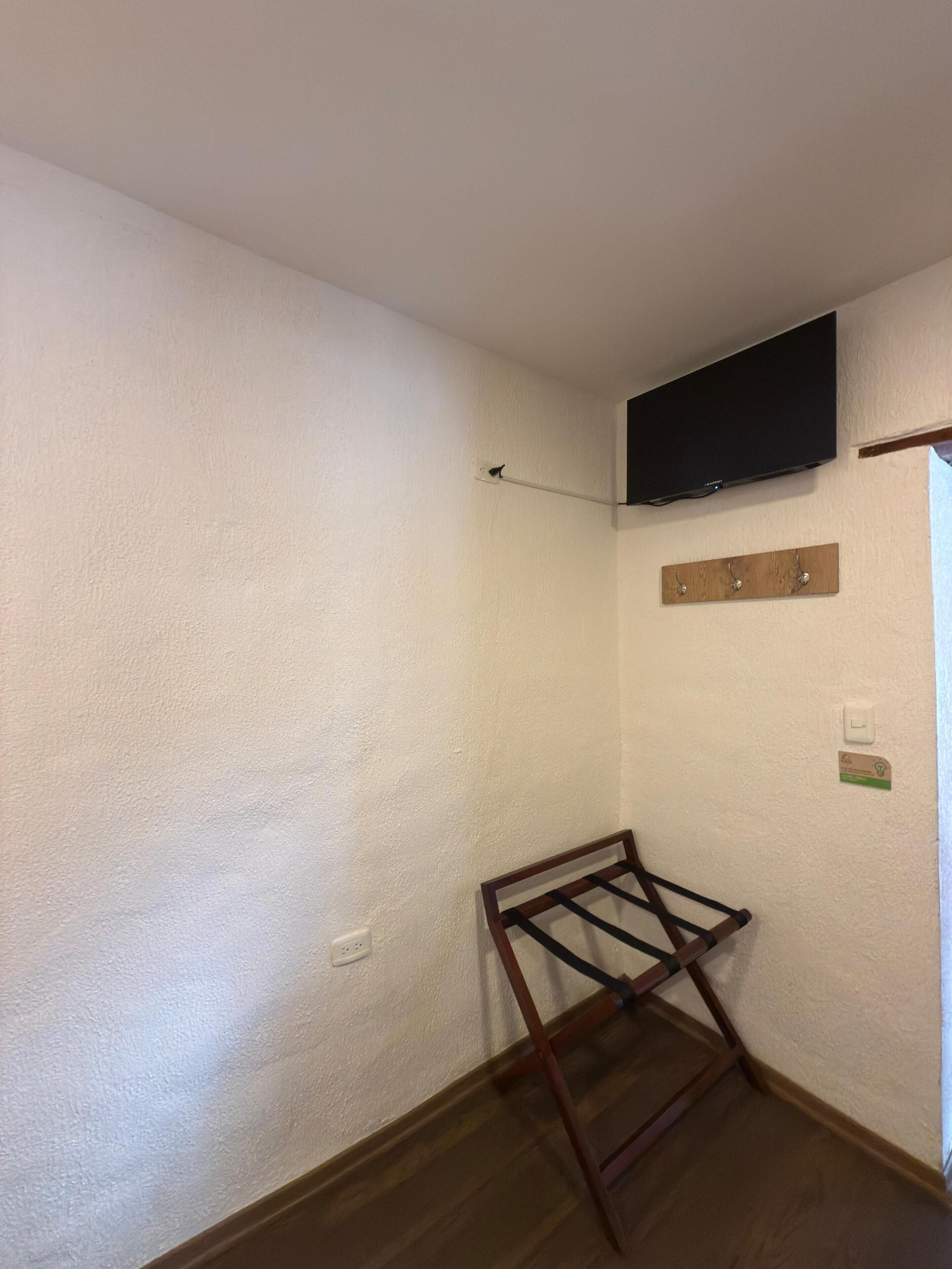 Una habitación con paredes blancas, suelo de madera, televisor montado, tendedero y un estante de madera con ganchos.