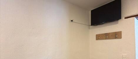 Una habitación con paredes blancas, suelo de madera, televisor montado, tendedero y un estante de madera con ganchos.
