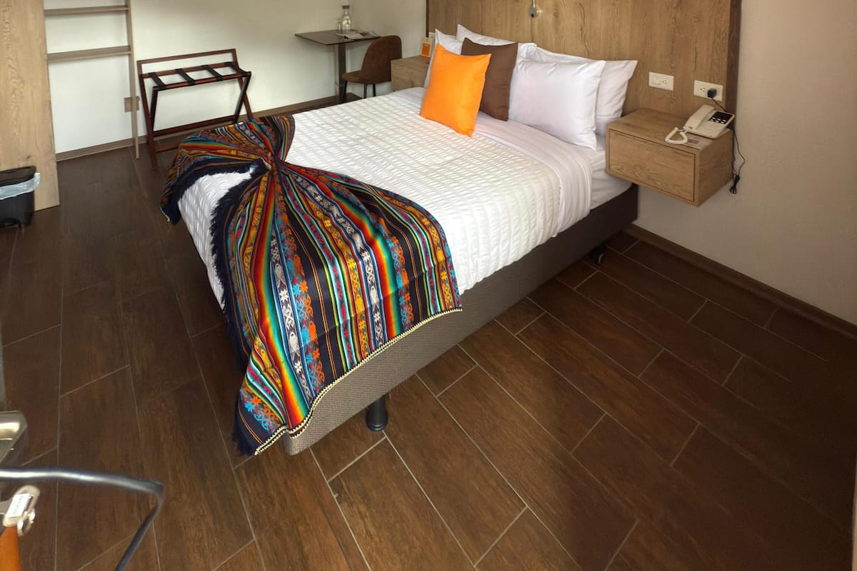 A bedroom with a bed, a bedside table with a phone, a chair, a desk, and a shelf.