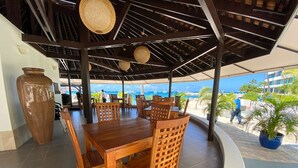 Un espace de restauration extérieur abrité, avec des tables et des chaises en bois, un grand vase et une vue sur la plage et l’océan.