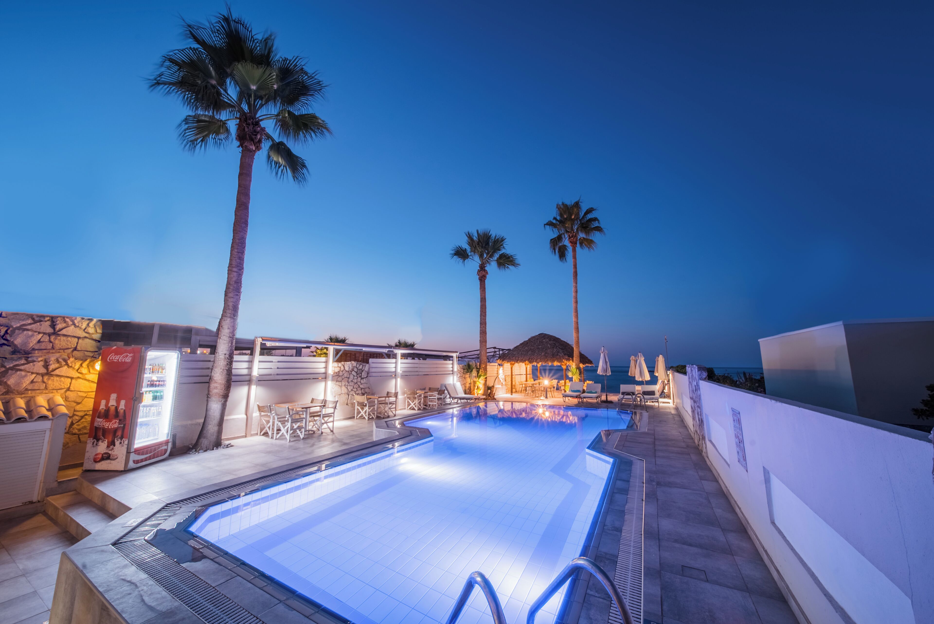 A swimming pool with lounge chairs and umbrellas, surrounded by palm trees and a stone wall.