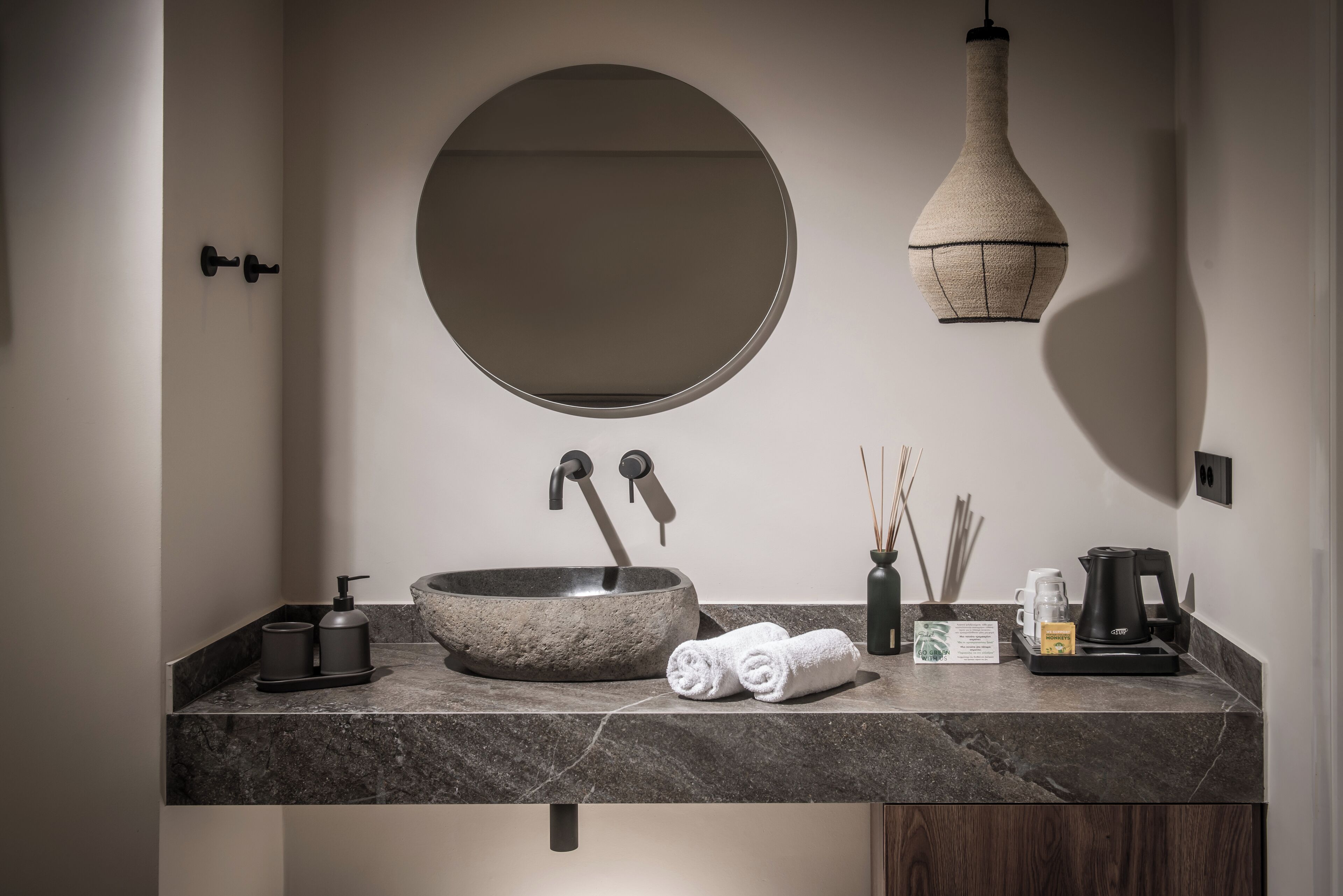 Un baño moderno con un espejo redondo, un lavamanos de piedra y una lámpara colgante.