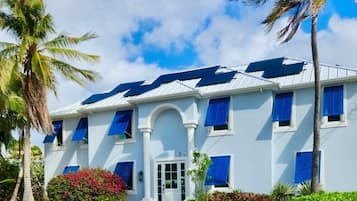 Un bâtiment à deux étages, doté d’une façade blanche et de volets bleus, entouré d’une végétation tropicale et de palmiers.