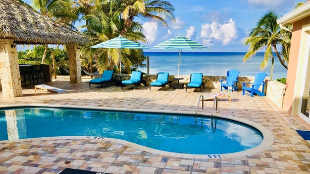 A pool area with lounge chairs and umbrellas, a stone wall, and a view of the ocean. - Reef Romance, Rum Point Oceanfront Villa with Private Pool and Gorgeous Beach! (Cayman Islands)