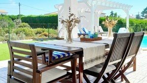 An outdoor dining area with a table and chairs, a pergola overhead, and a view of a swimming pool. - Villa Sonho do Mar - Villa with Saltwater Pool (Vale da Telha)