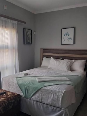 Una cama bien hecha con cabecera de madera, una mesita de noche con control remoto y una ventana con cortinas traslúcidas.