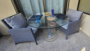 Une table ronde en verre, quatre chaises en osier gris, un plateau en verre et un petit tapis.