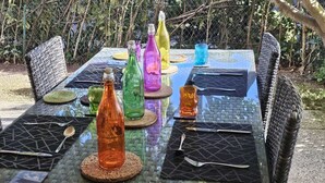 Une table de jardin, dressée pour un repas en plein air, agrémentée de bouteilles en verre colorées, d’assiettes et de couverts.