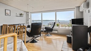 A modern living room with a large window overlooking a body of water, a black leather chair, a wooden table, and a black cabinet.