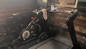 Ein stationäres Fahrrad, ein Laufband und ein Ventilator in einem Fitnessraum mit Holzwänden und einem Fenster.