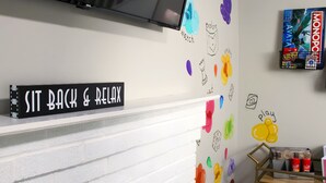 Una stanza con un cartello che riporta la scritta "SIT BACK & RELAX" su un muro di mattoni bianchi. Si vedono disegni colorati e un gioco da tavolo Monopoly appesi al muro.