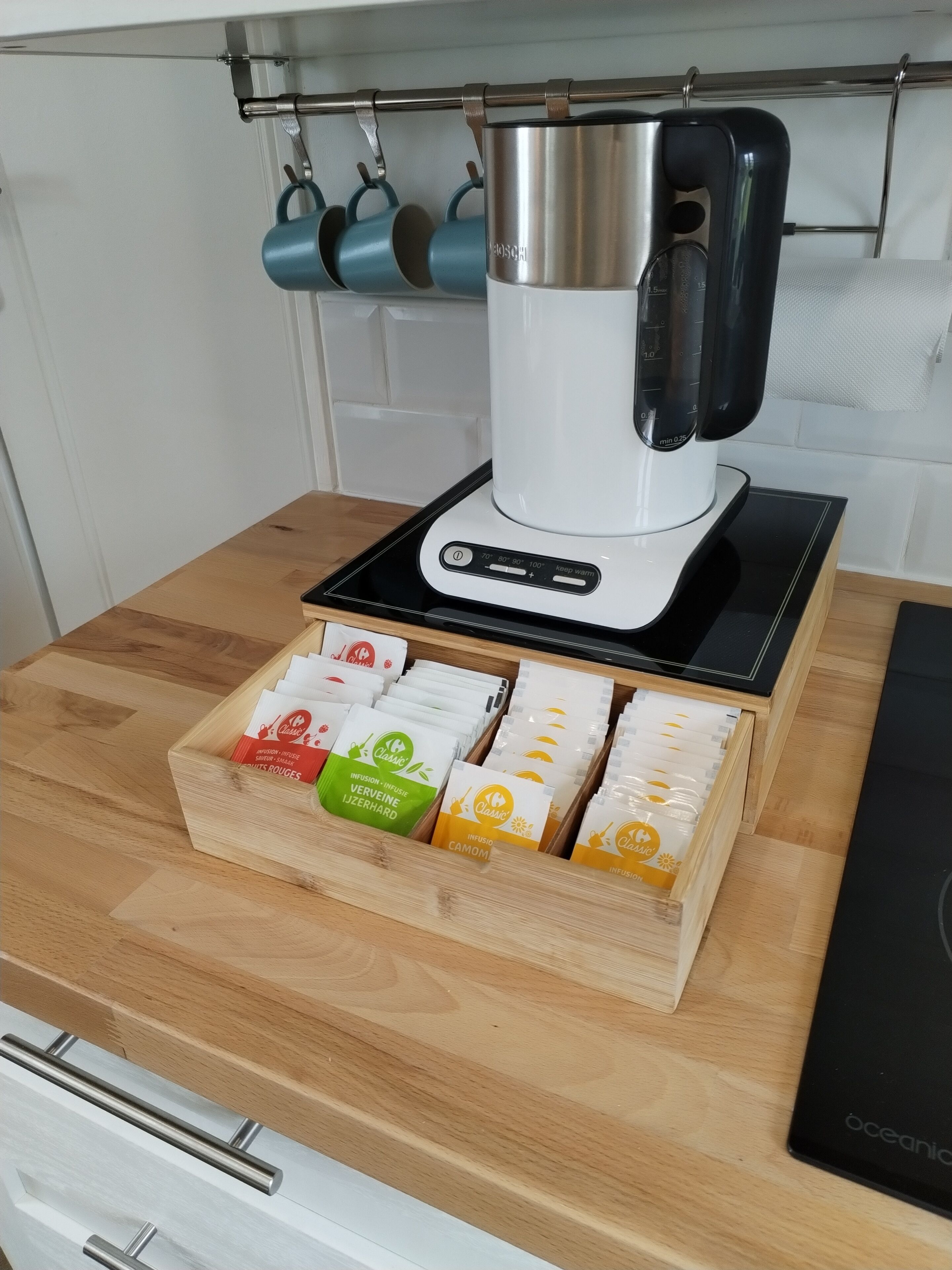 A kitchen with a coffee maker on a black countertop, a wooden box with tea bags, and a hanging rack with cups.