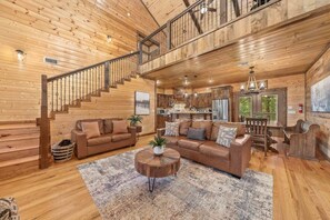 A spacious living area with a wooden staircase, a sofa set, a coffee table, and a view of the kitchen.