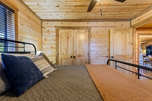 A wooden cabin interior with a bed, a ceiling fan, and a view into the kitchen area.