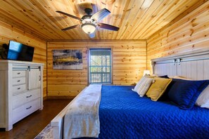 A wooden cabin bedroom with a bed, a ceiling fan, a television, and a window.