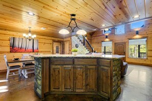Interior de una cabaña de madera con una isla central, zona de comedor y una escalera que sube a un nivel superior.
