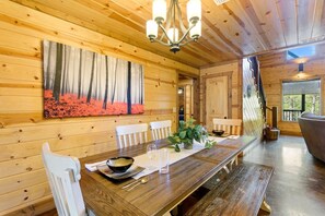 A wooden interior with a dining table, chairs, a chandelier, and a painting on the wall.