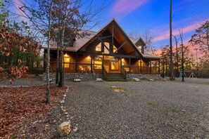 A log cabin with a stone foundation, a porch, and a gravel driveway.