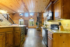 Interior de una cabaña de madera con cocina, comedor y una escalera que sube a un nivel superior.