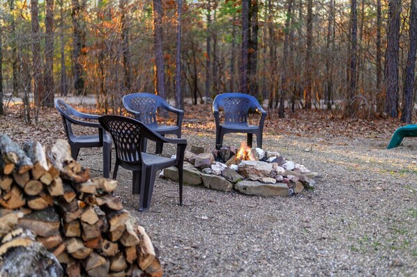 A forest setting with a fire pit and chairs.