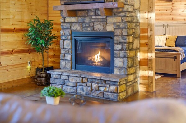A stone fireplace with a visible fire, a wooden bed, and a potted plant in a log cabin interior.