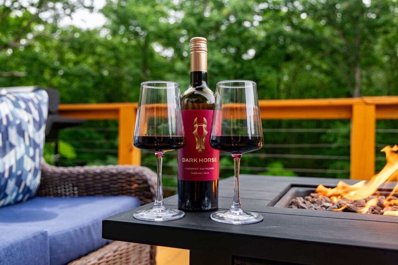 A bottle of Dark Horse Cabernet Sauvignon on a table with two glasses of wine, set outdoors with a wooden deck and greenery in the background.