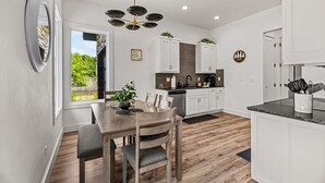 Una cocina moderna con armarios blancos, una mesa de comedor con sillas, una lámpara de araña y vistas a zonas verdes.