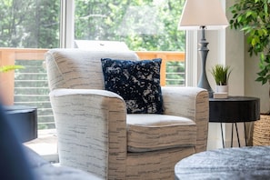 Une chaise recouverte d’un tissu à motifs, un coussin bleu foncé et blanc, une lampe et une vue sur la verdure à l’extérieur.