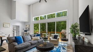 A spacious living room with a large sofa, a coffee table, and a television. There is a staircase leading to an upper level, and large windows providing a view of the outdoors. - Modern Chalet Near Back 40 Trails Hot Tub & Forest Views (Bella Vista)