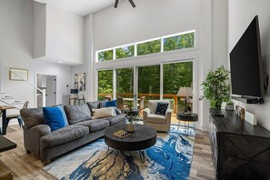 A spacious living room with a large sofa, a coffee table, and a television. There is a staircase leading to an upper level, and large windows providing a view of the outdoors. - Modern Chalet Near Back 40 Trails Hot Tub & Forest Views (Bella Vista)
