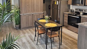 Eine moderne Küche mit Holzausstattung, einem Edelstahlkühlschrank, einer Mikrowelle und einem Herd. Ein Esstisch mit Stühlen und ein Blumentopf sind ebenfalls vorhanden.