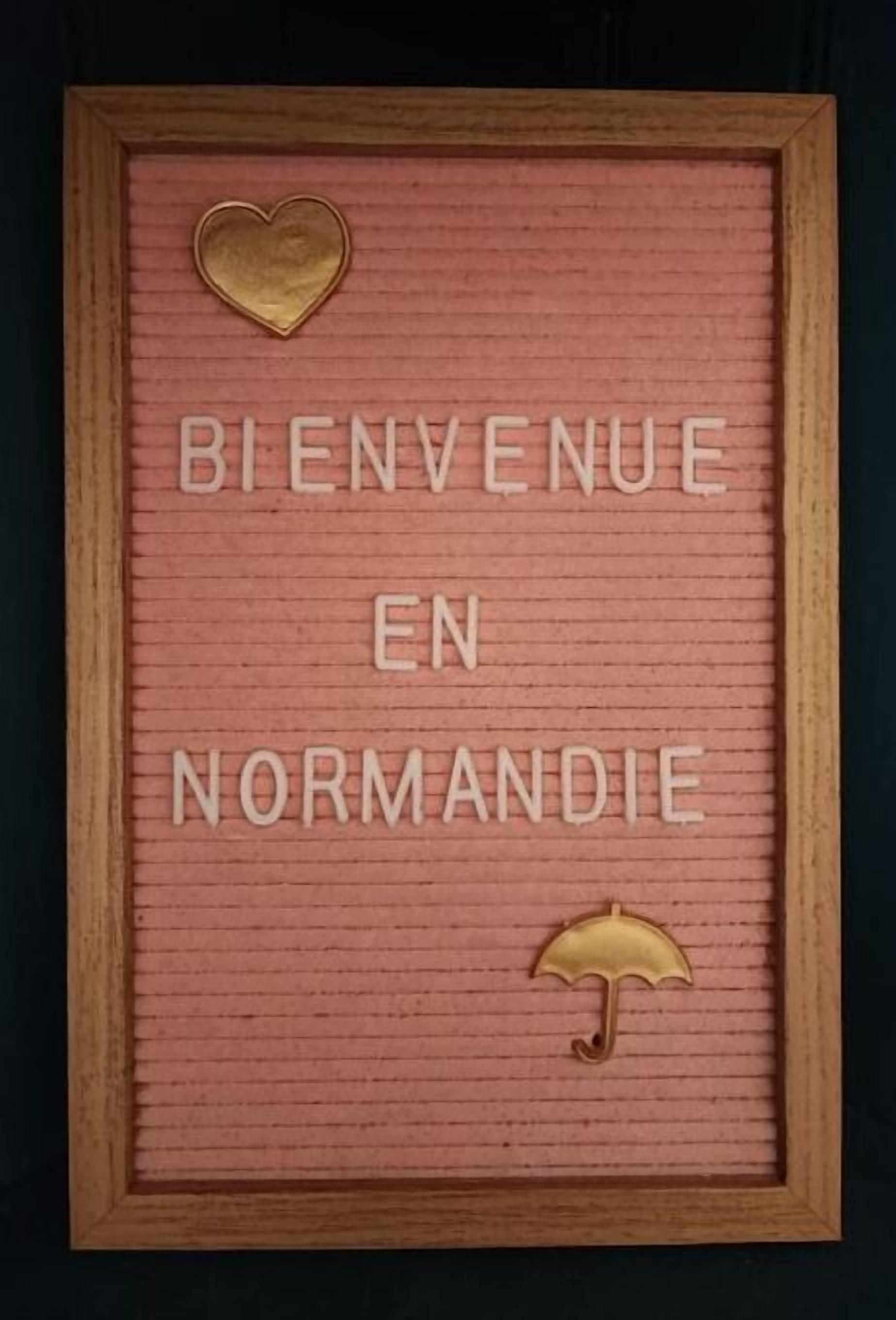 A framed welcome sign with the text "Bienvenue en Normandie" and decorative elements like a heart and an umbrella.