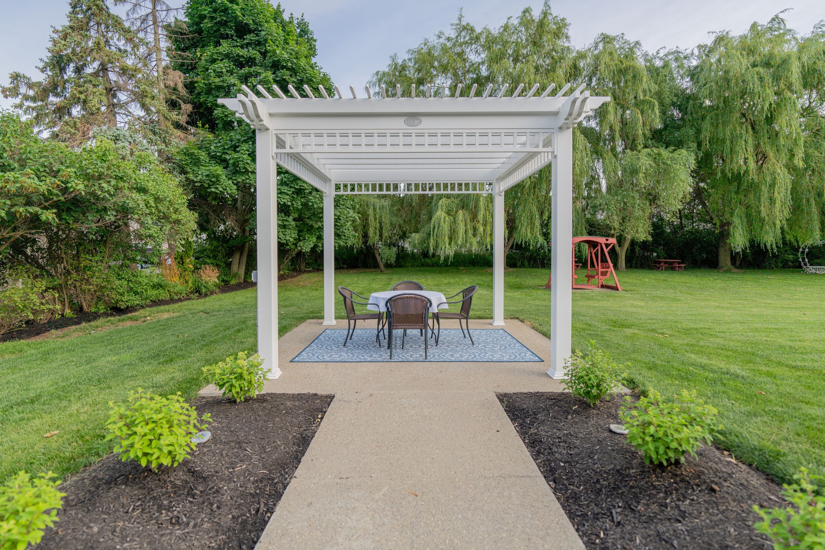 Un jardin agrémenté d’une pergola blanche, d’une table avec des chaises et d’une balançoire.