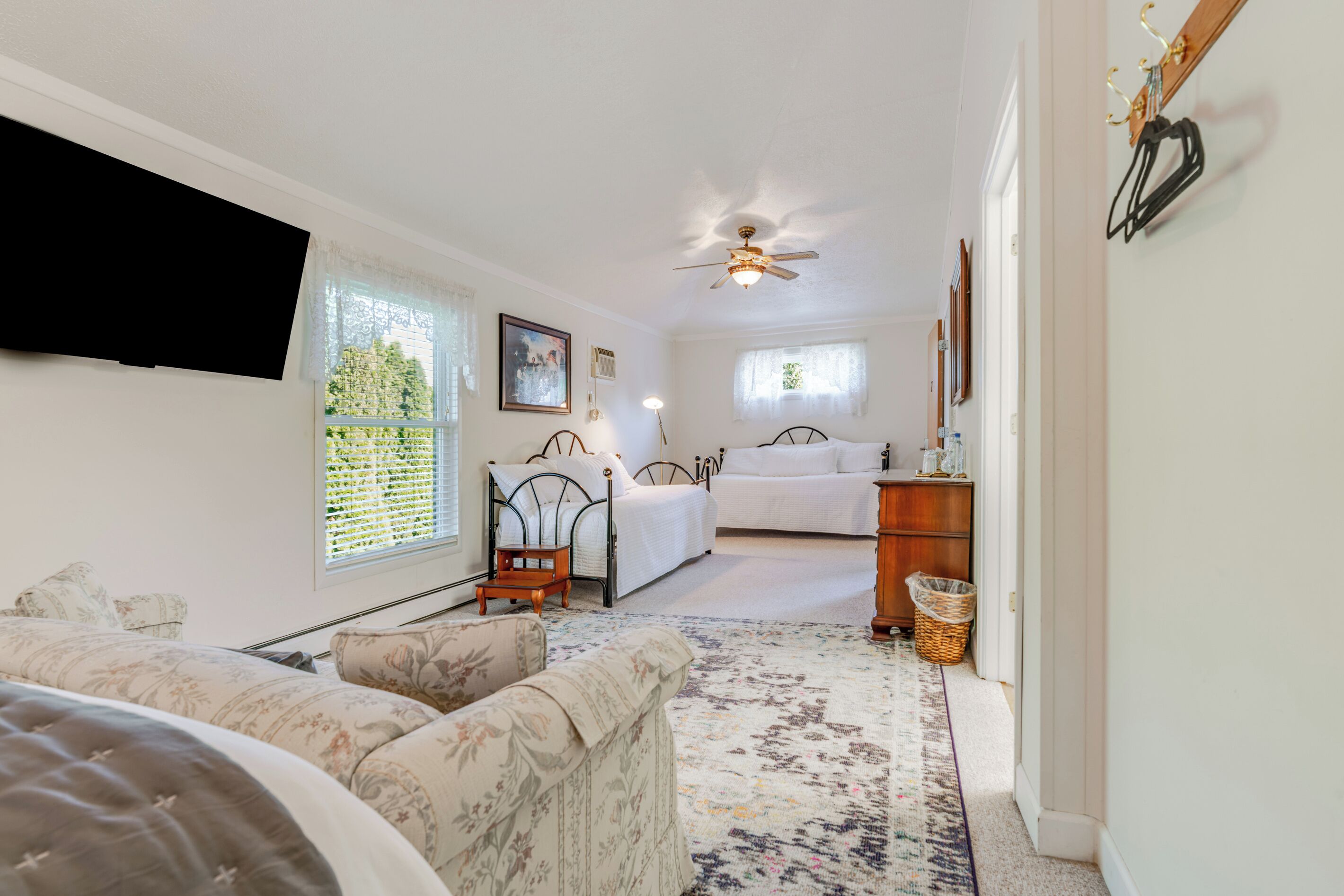 A bedroom with a bed, bedside table, and a nightstand. There is a window with blinds, a ceiling fan, and a wall-mounted TV.