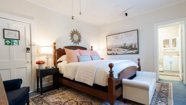 A bedroom with a four-poster bed, a nightstand with a lamp, a chair, a patterned rug, and a painting on the wall.