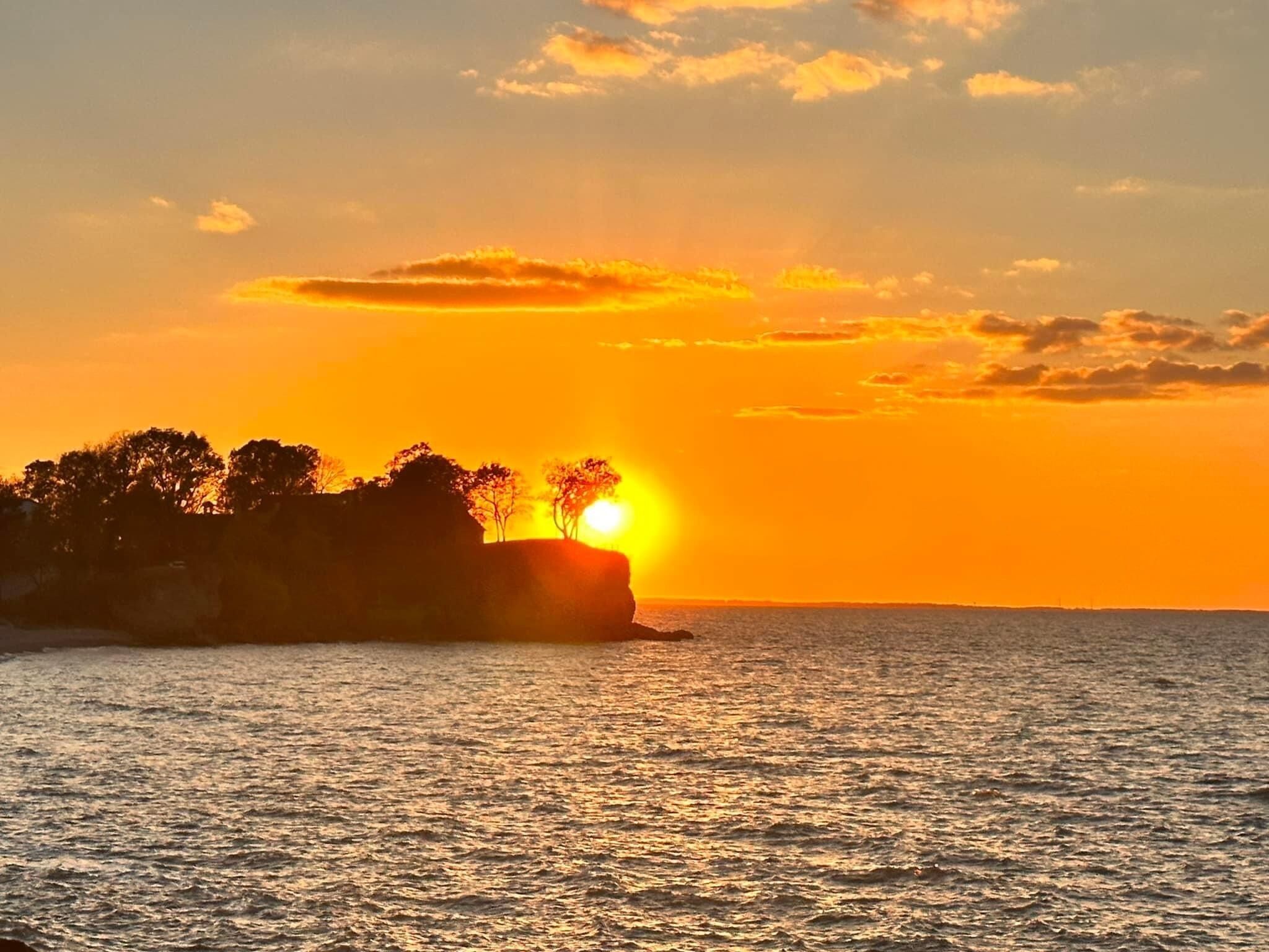 Un coucher de soleil sur une étendue d’eau, avec en silhouette des arbres et une falaise.
