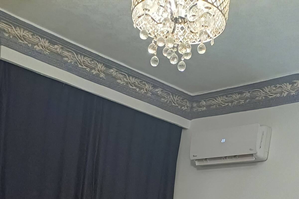 Een slaapkamer met een groot bed, een nachtkastje, een kroonluchter en een raam met gordijnen.