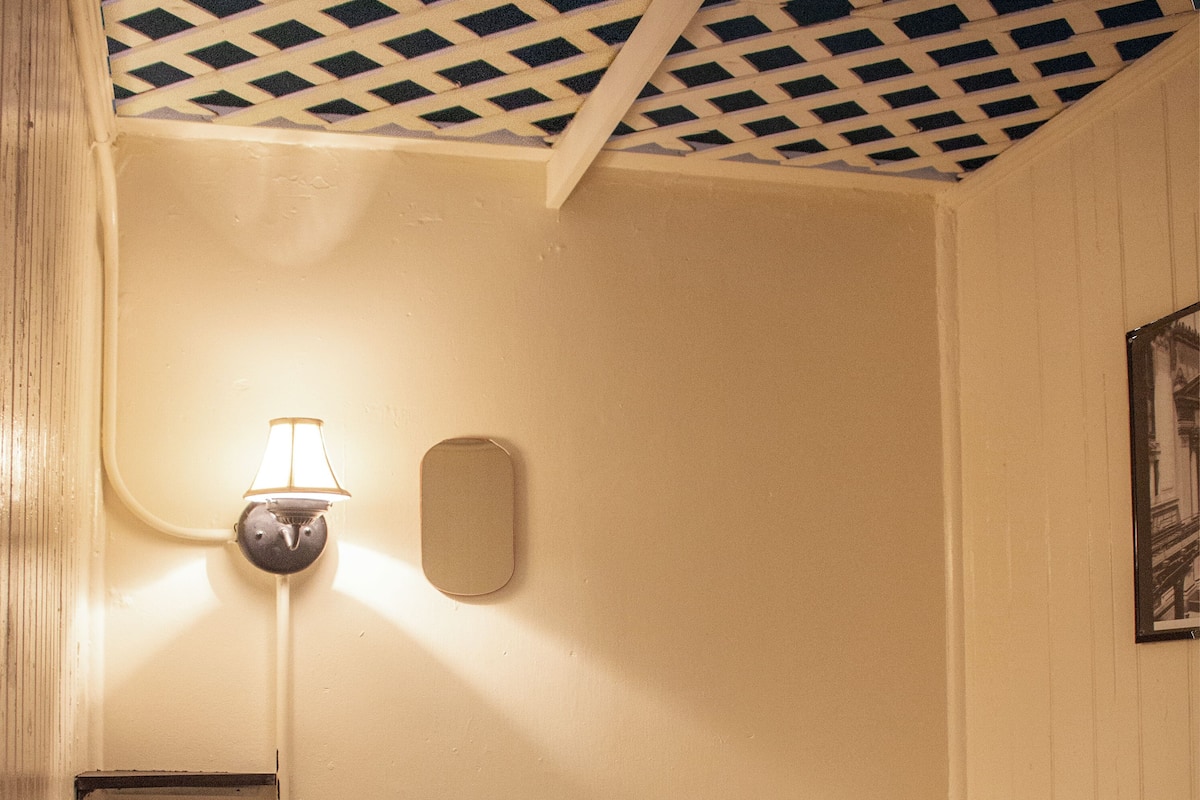 A small, single-bed room with a white bed, a nightstand, a wall-mounted lamp, and a power outlet.