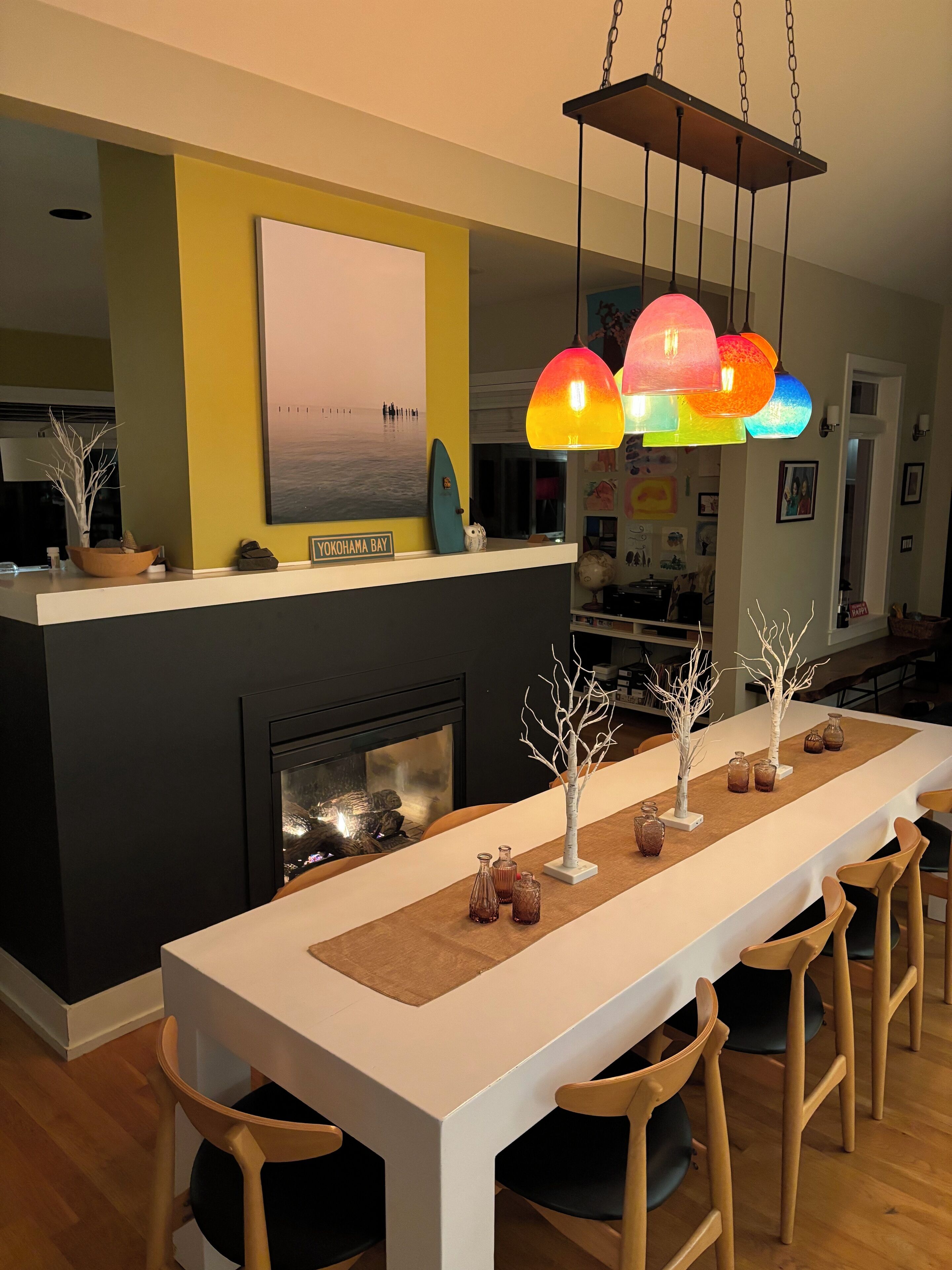 A dining area with a long table, chairs, a fireplace, and a colorful pendant light.
