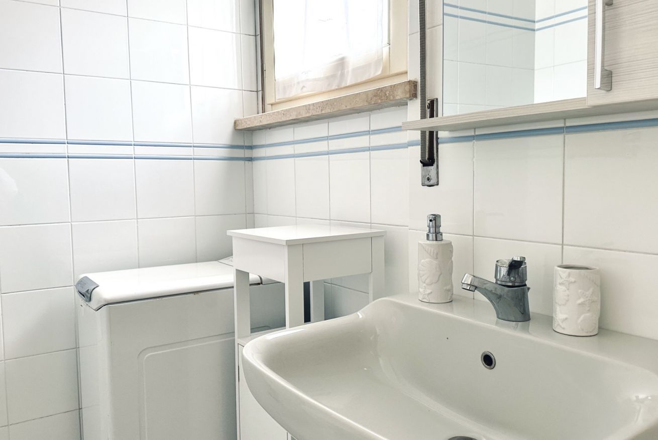 A bathroom with white tiles, a sink, a mirror, and a window with a curtain.