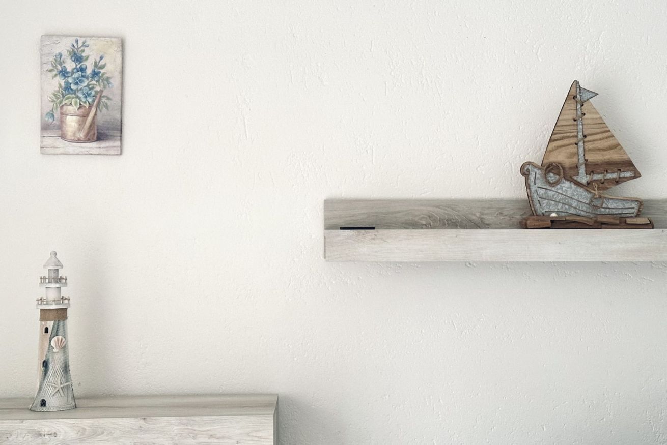 A shelf with a wooden sailboat model, a lighthouse model, and a framed painting of a potted plant.