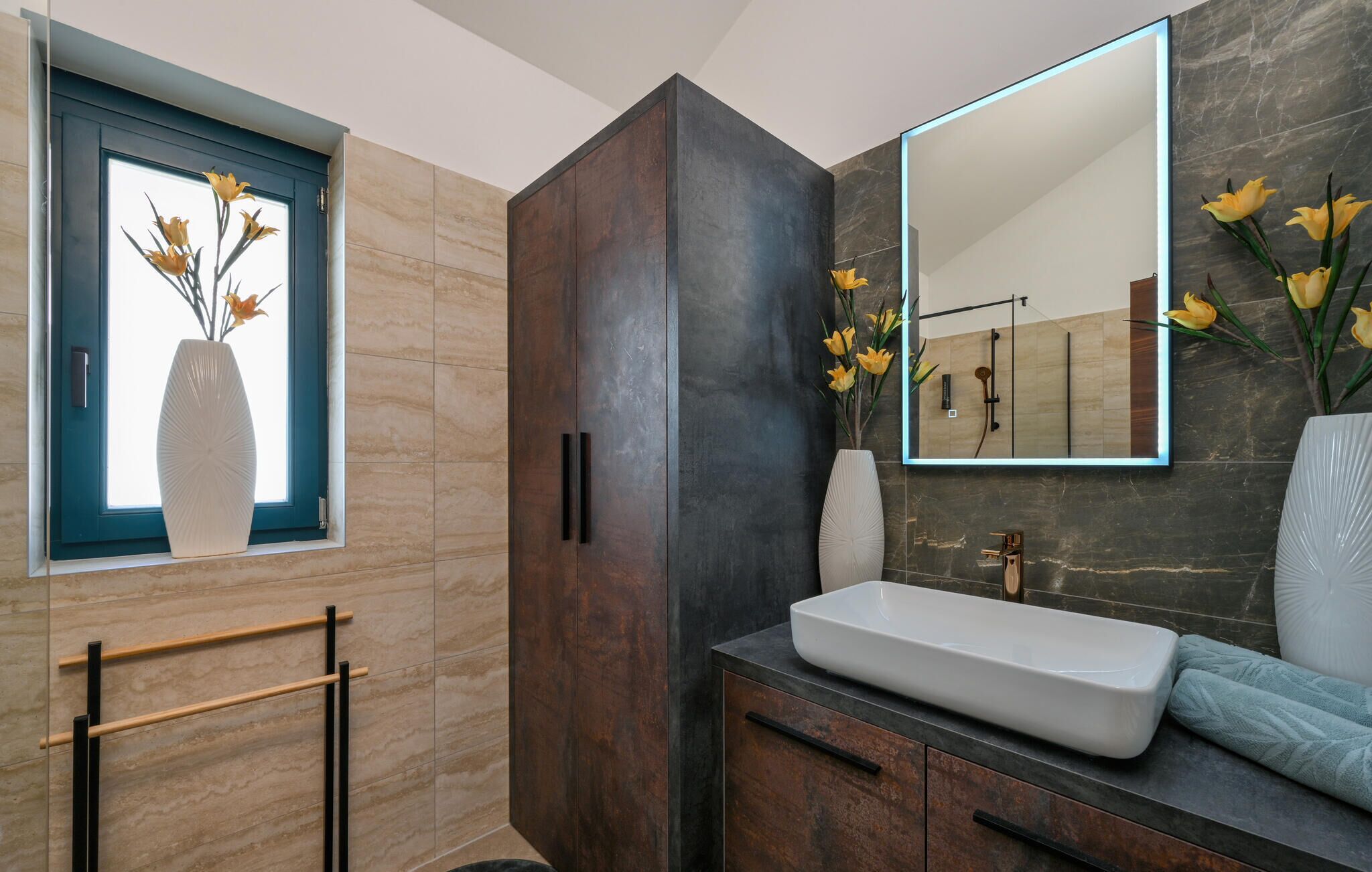 A modern bathroom with a large mirror, a white sink, and a window with a vase of flowers.