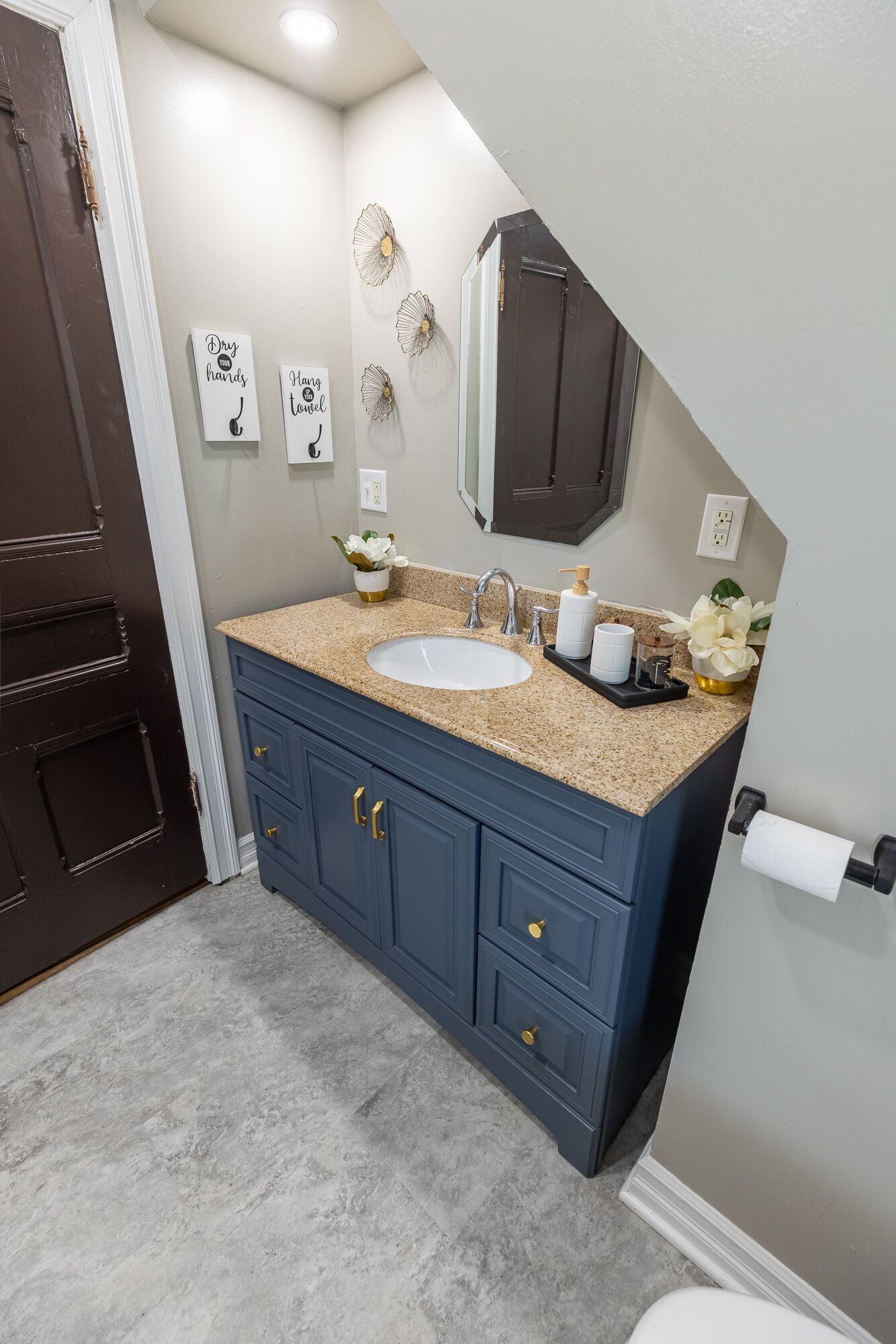 Une salle de bain avec un meuble-lavabo bleu, un lavabo blanc et un miroir au-dessus. On y trouve également des éléments décoratifs sur les murs et un porte-rouleau de papier toilette.