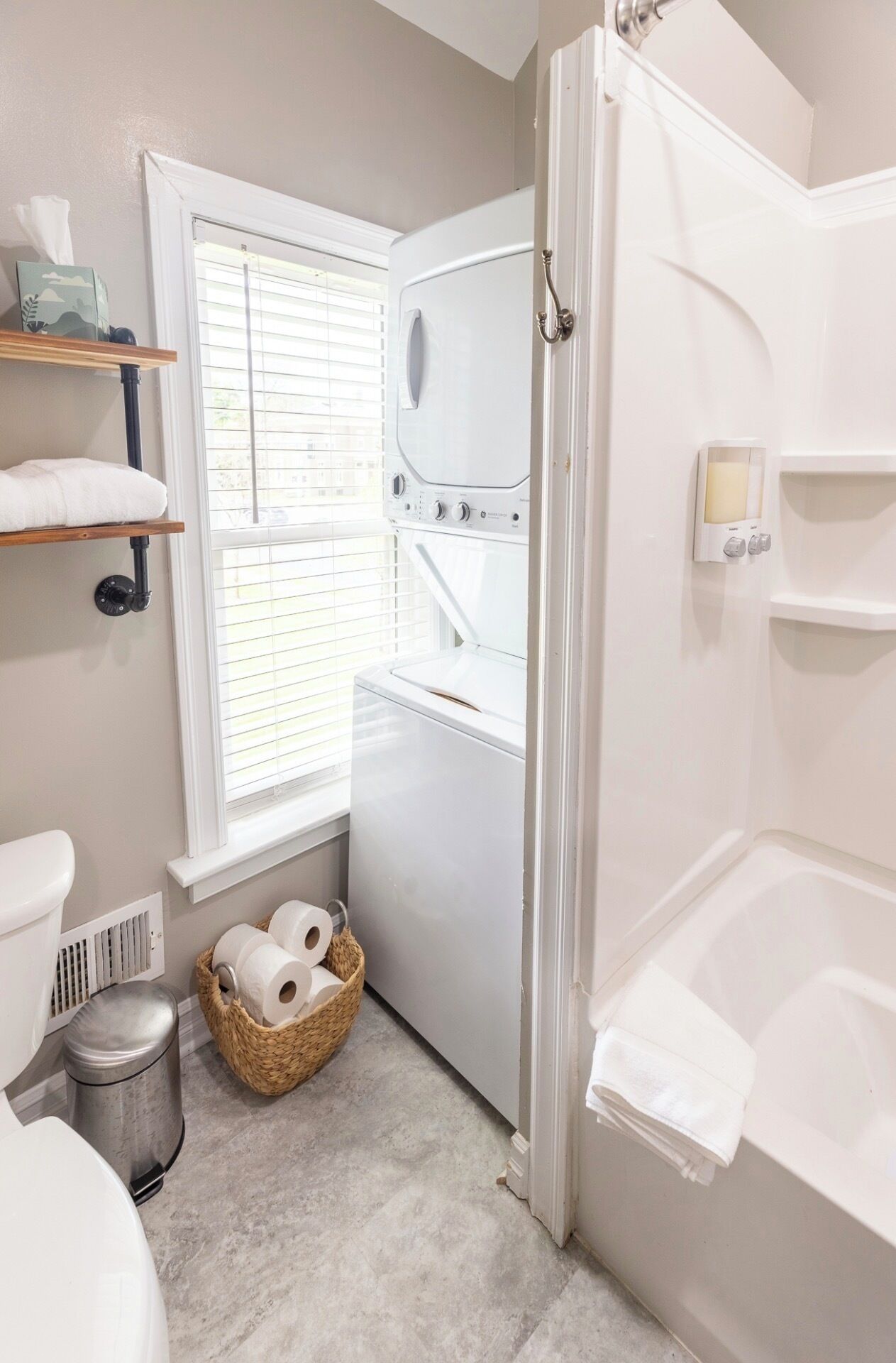 Ein Badezimmer mit Toilette, Waschmaschine, Dusche und einem Fenster mit Jalousien.